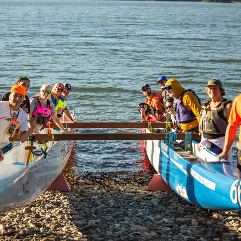 A group of people in colorful vests stand by two canoes on a rocky shore, with a calm body of water stretching out behind them.