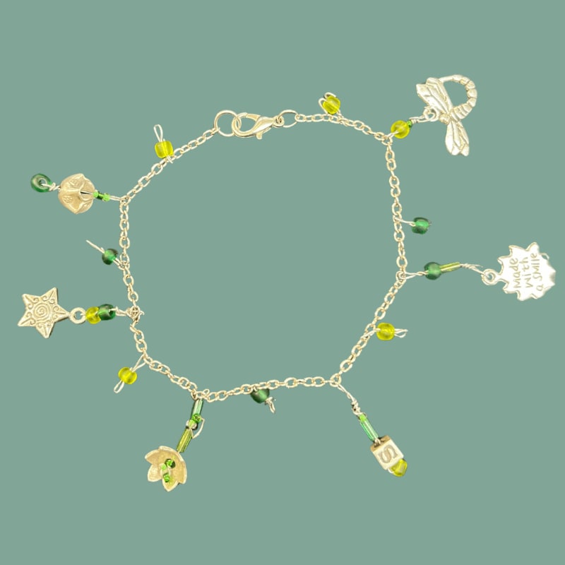A delicate silver bracelet adorned with green beads and charms like a star, flower, and dragonfly hangs against a muted green background.