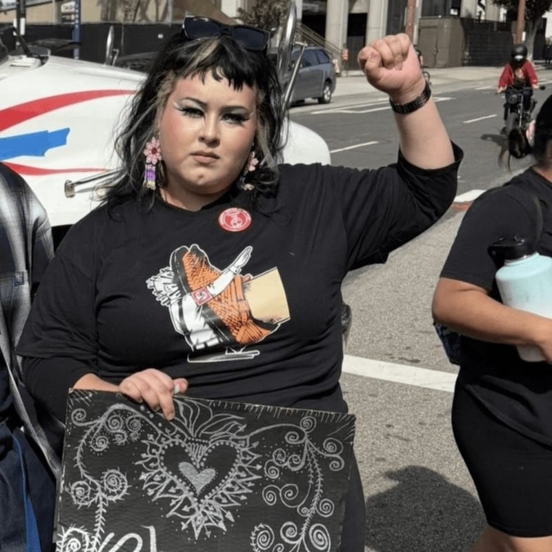 A woman with dark hair and bold makeup raises her fist in the foreground, holding a sign, while a truck and a person on a scooter are visible in the blurred background.