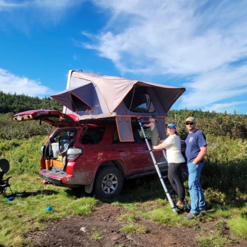A couple stands by their red SUV with an open rooftop tent in a grassy field, with trees and a blue sky in the background.