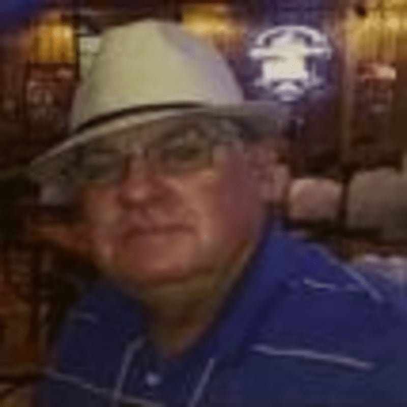 A man in a white fedora and blue shirt is in the foreground, with a dimly lit bar or restaurant in the background.