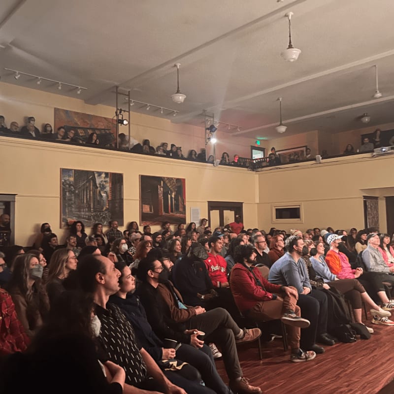 A diverse audience fills a theater, seated on the main floor and in the balcony, with artwork adorning the walls.