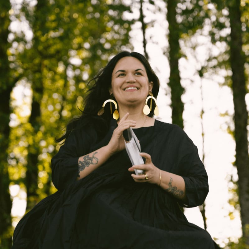 A smiling woman in a black dress holds a crystal in front of a blurred background of trees.