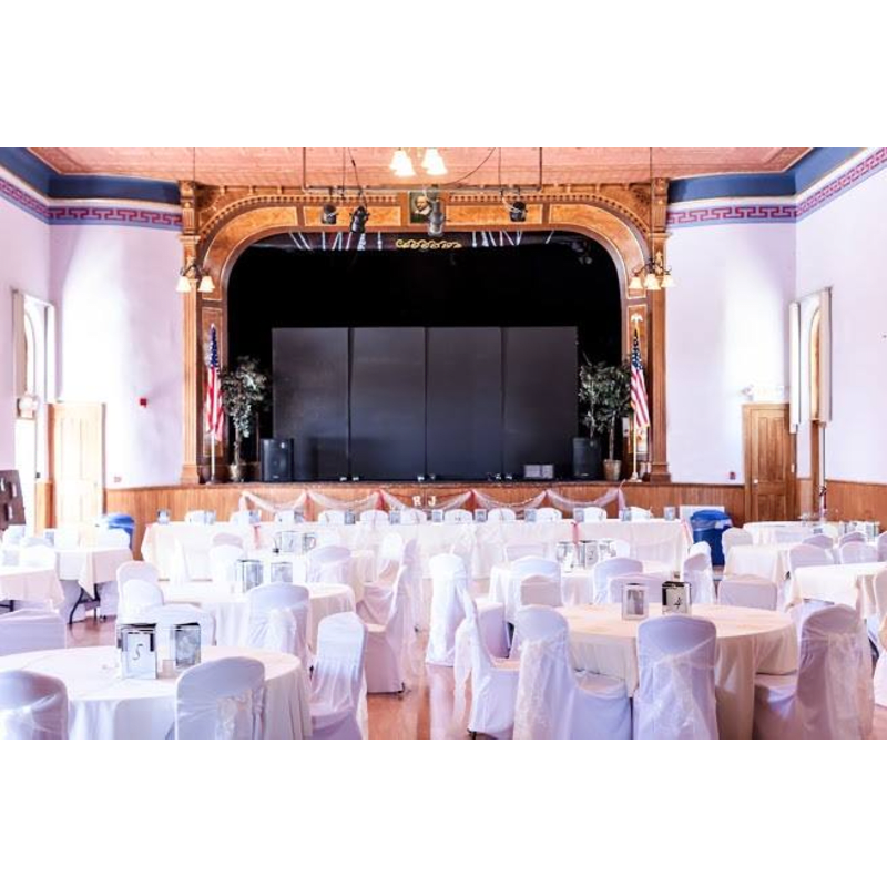 White-clothed round tables fill the foreground of a grand hall with a stage and a large black screen in the background.
