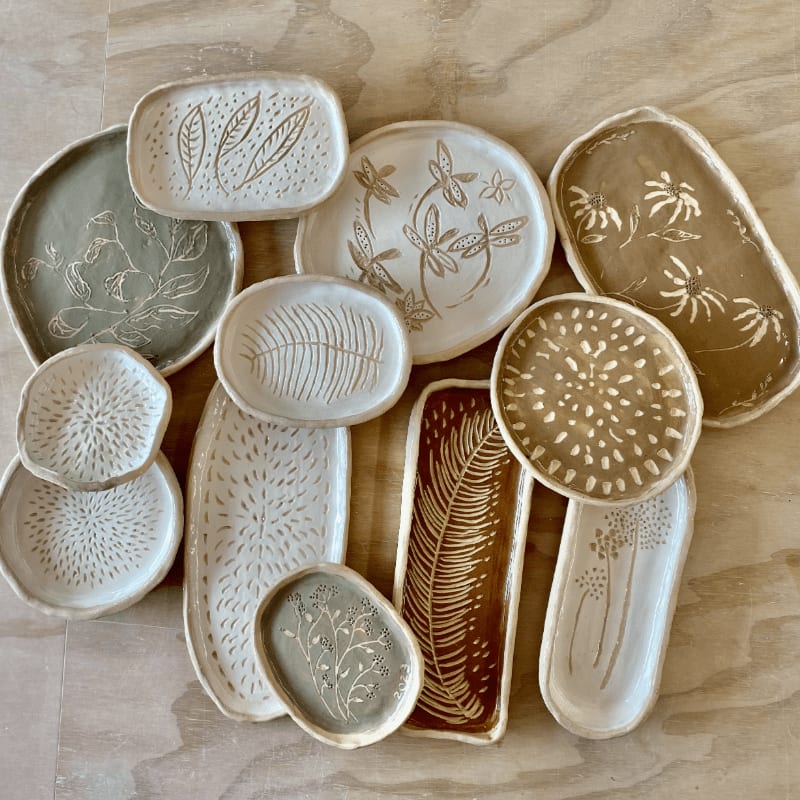A collection of handmade ceramic dishes with botanical designs are arranged on a light wooden surface.