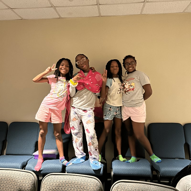 Four children in pajamas stand on chairs in front of a plain wall, with rows of blue chairs behind them.