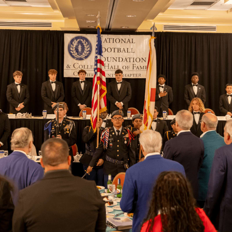 In the foreground, people in formal attire stand at tables, while in the background, young men in military-style uniforms stand behind a banner for the National Football Foundation & College Football Hall of Fame.