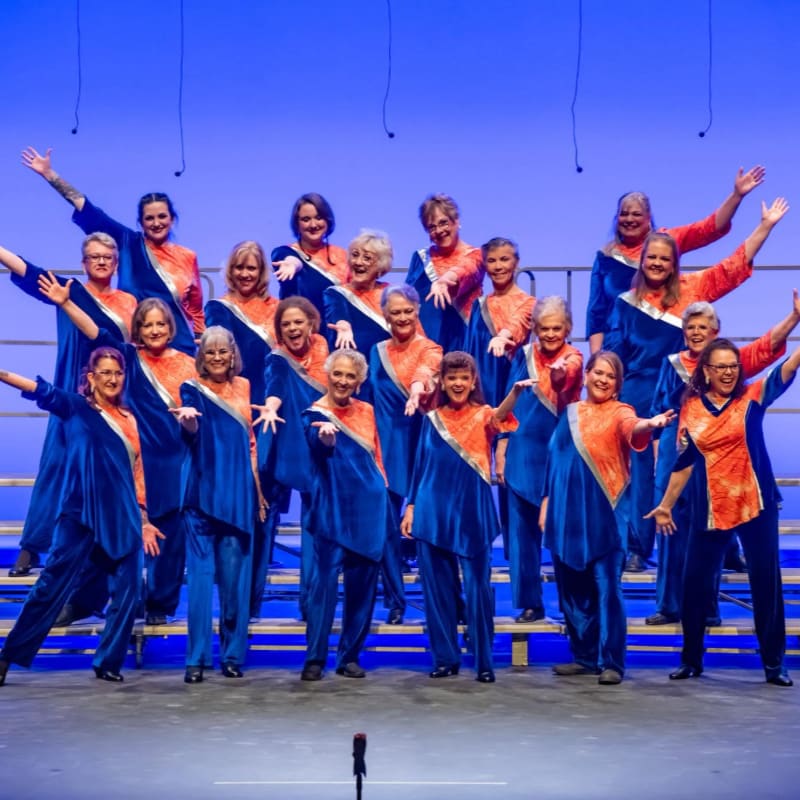A choir of women in matching blue and orange outfits stands on risers against a blue backdrop, with some members raising their arms in a performance.
