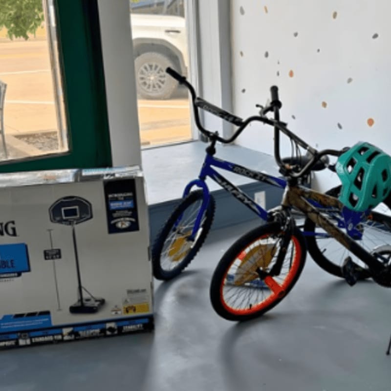 A large box for a basketball hoop sits in the foreground next to two BMX bikes, one blue and one camouflage, with a teal helmet on the handlebars, all in front of a window with a white SUV visible outside and a wall decorated with colorful polka dots.