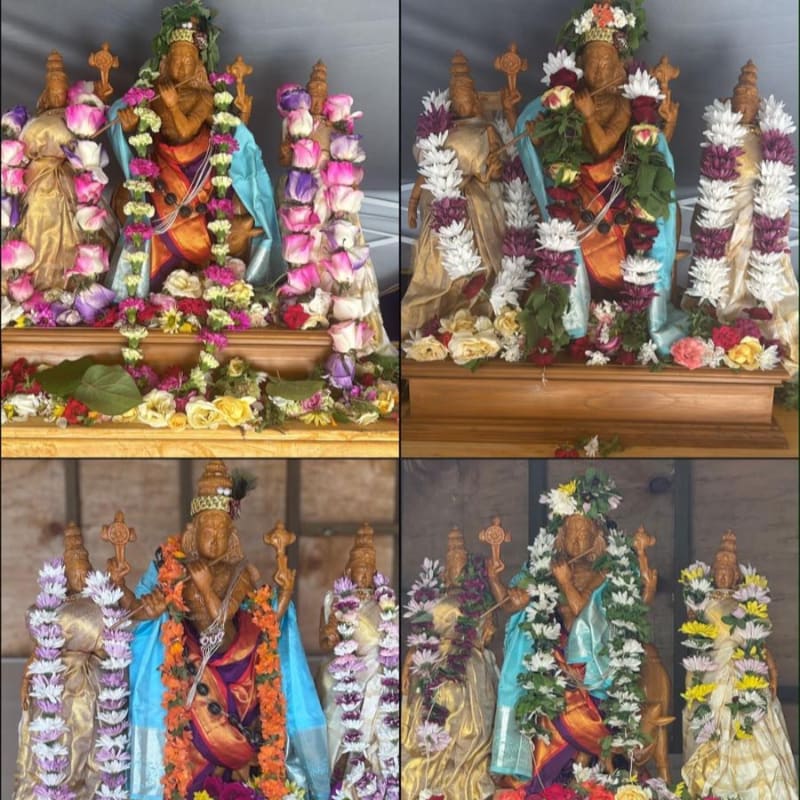 Four ornate statues of deities, adorned with garlands of colorful flowers and draped in vibrant fabrics, stand on wooden pedestals against a muted, textured background.
