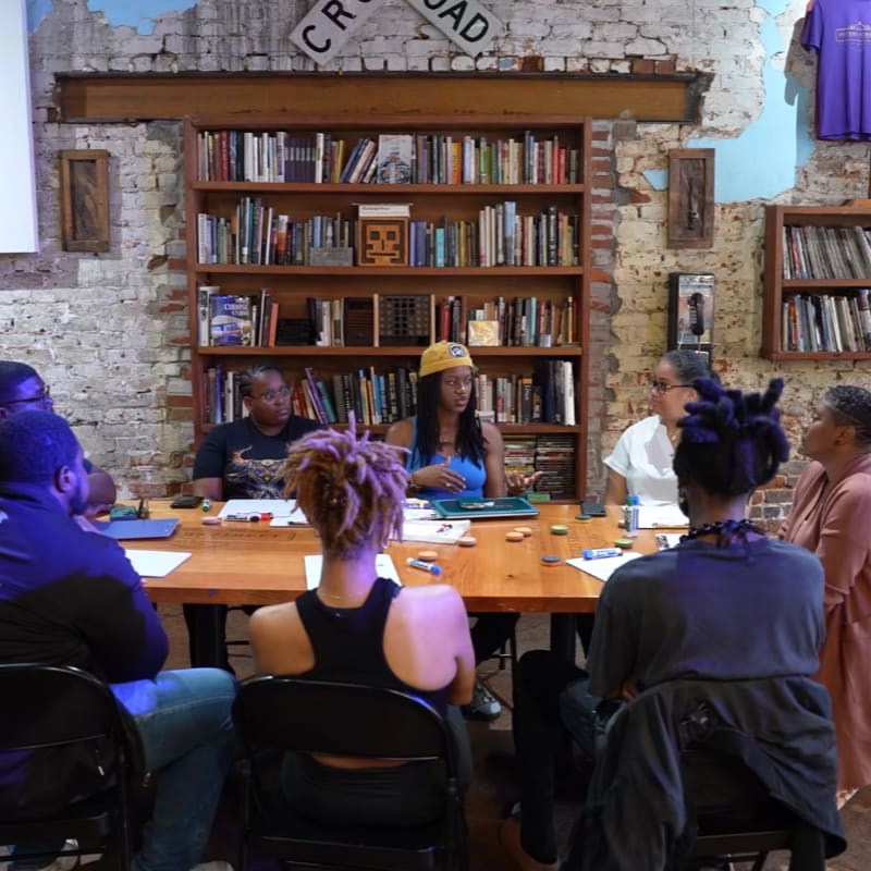 A group of people sit around a wooden table in a room with bookshelves and exposed brick walls.