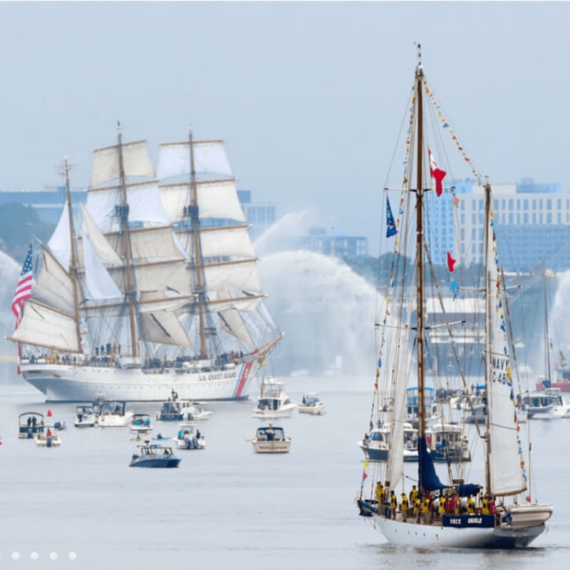 A white sailboat with colorful flags in the foreground sails past a large white US Coast Guard barque with full sails, surrounded by smaller boats and water cannons in the hazy background.