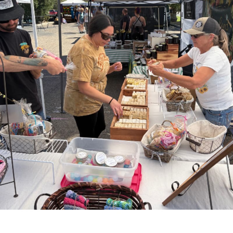 People are browsing and buying handmade goods at an outdoor market with stalls and tents in the background.