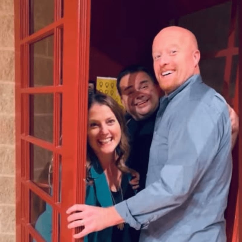 Three smiling people, two men and a woman, are framed by a red doorway, with a textured wall visible in the background.