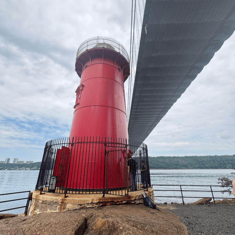 A bright red lighthouse stands on a rocky shore in the foreground, with a large suspension bridge overhead and a city skyline visible across the water in the background.