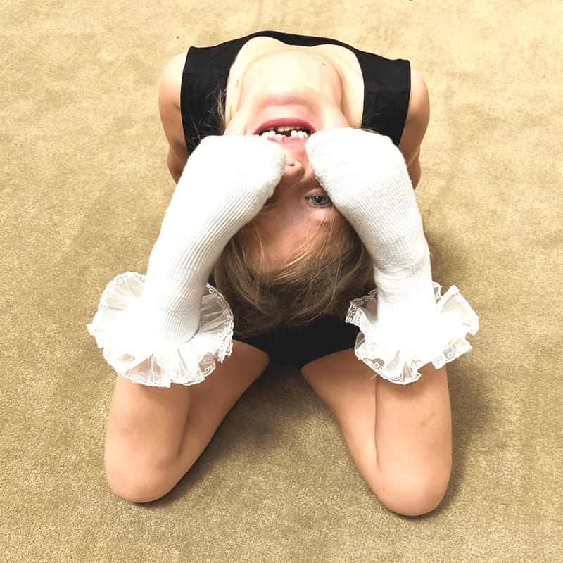 A young girl in a black leotard and white ruffled socks is kneeling on a beige carpet, with her head tilted back and her feet in the air.