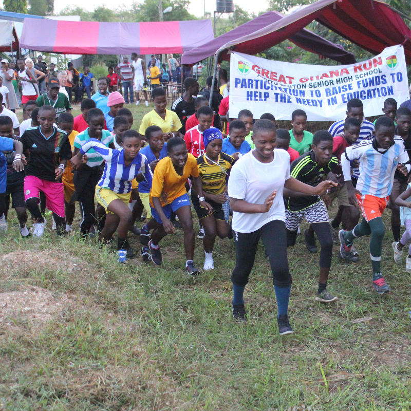 A group of students in athletic wear are running a race in the foreground, with tents and spectators visible in the background.