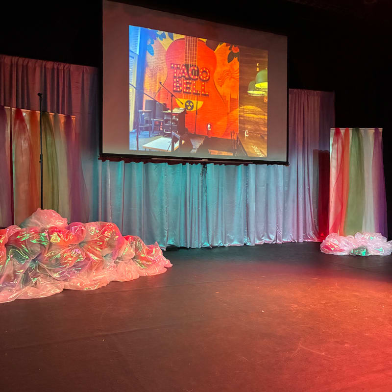 A stage is set with colorful fabric draped on either side of a large projection screen displaying a guitar with "Taco Bell" written on it, while iridescent fabric is piled in the foreground.
