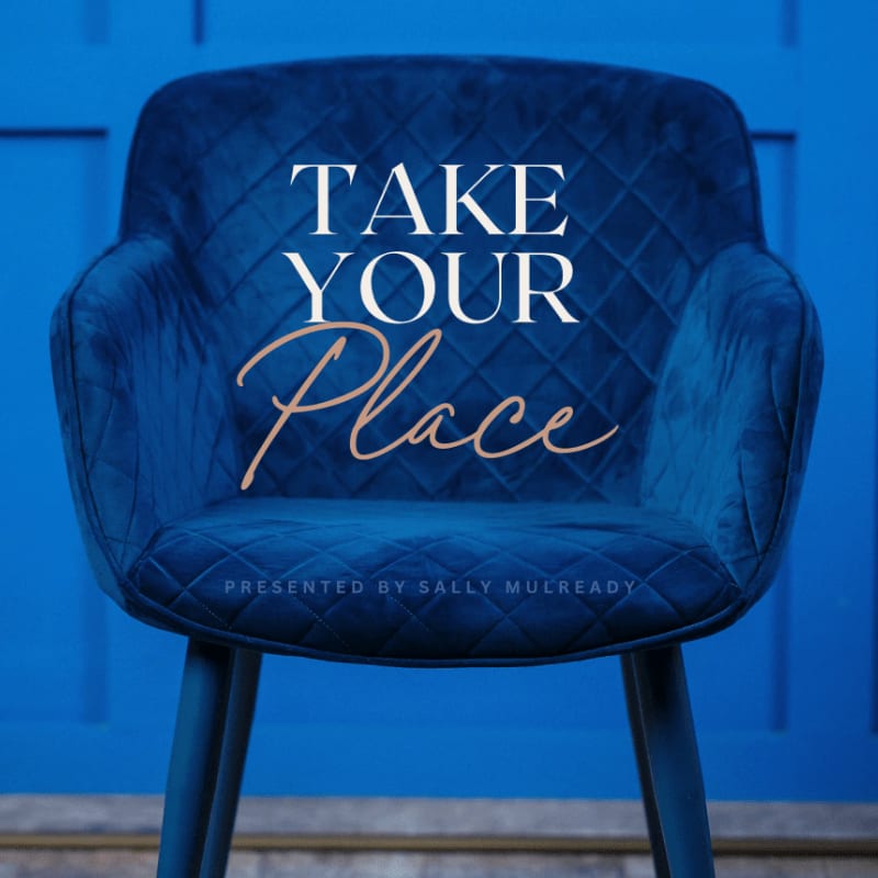 A plush blue armchair with the words "TAKE YOUR PLACE" and "PRESENTED BY SALLY MULREADY" emblazoned on it sits in front of a vibrant blue paneled wall.