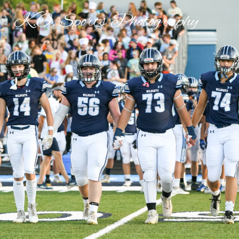 Four football players in navy blue uniforms with white pants walk onto a field with a crowd in the background.