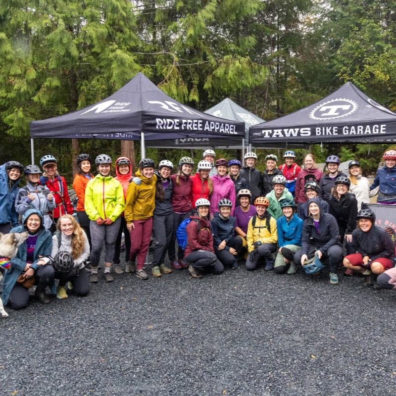 A group of women in cycling gear pose for a photo in front of tents, with a wooded area in the background.
