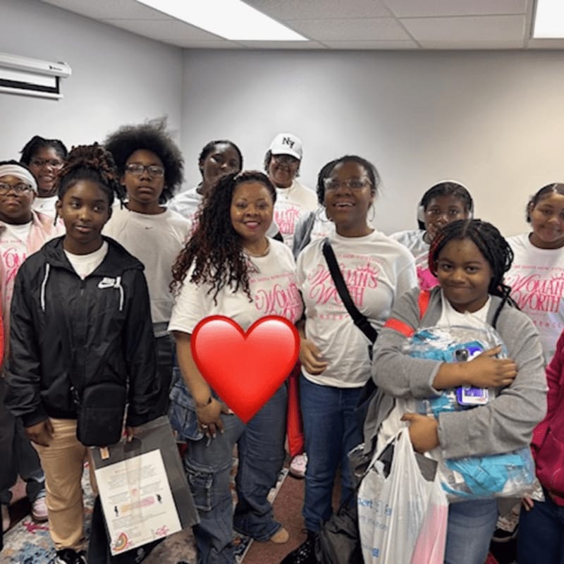 A group of young women in pink shirts stand in a room with a large red heart overlaying the center.