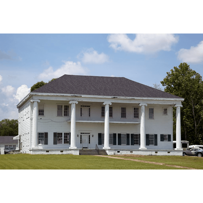 A grand white building with large columns stands on a green lawn, with trees and a cloudy blue sky in the background.