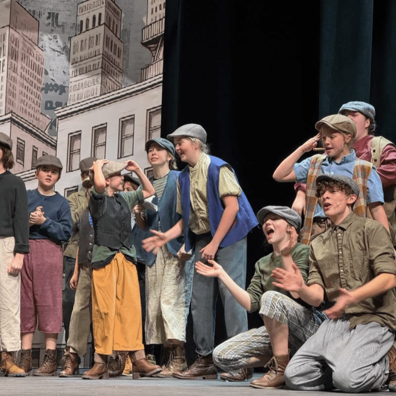 A group of children in period costumes stand on a stage with a painted cityscape backdrop.