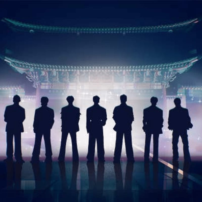 Silhouetted figures stand in the foreground before a brightly lit, ornate traditional building in the background.