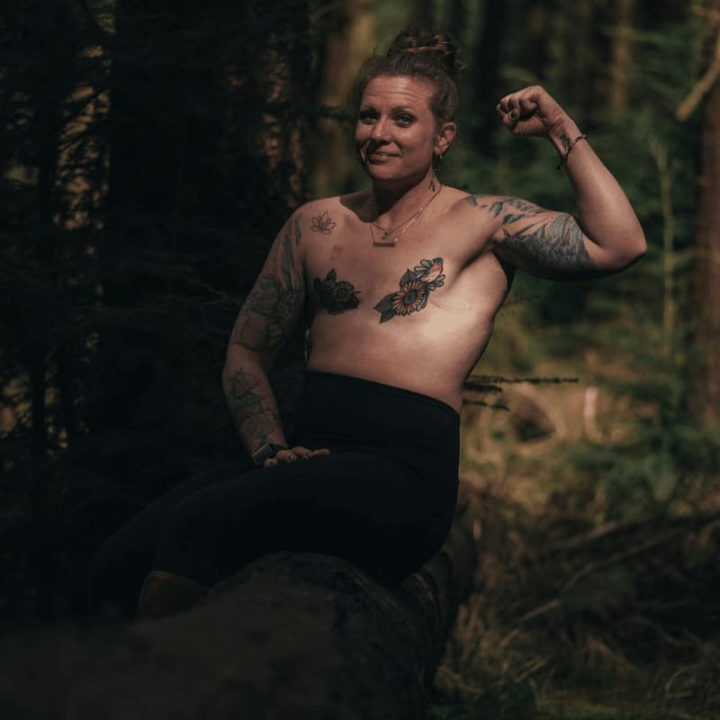 A tattooed woman with a raised arm sits on a fallen log in a dark forest.