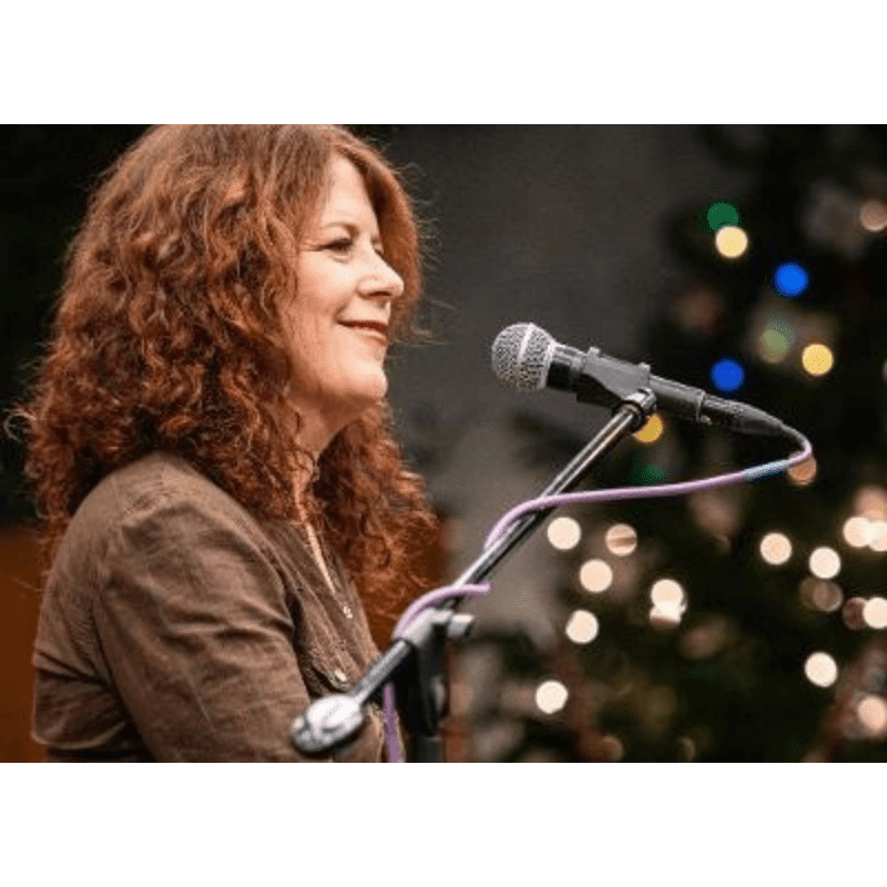A woman with curly red hair sings into a microphone in the foreground, with a blurred background of bokeh lights and a Christmas tree.