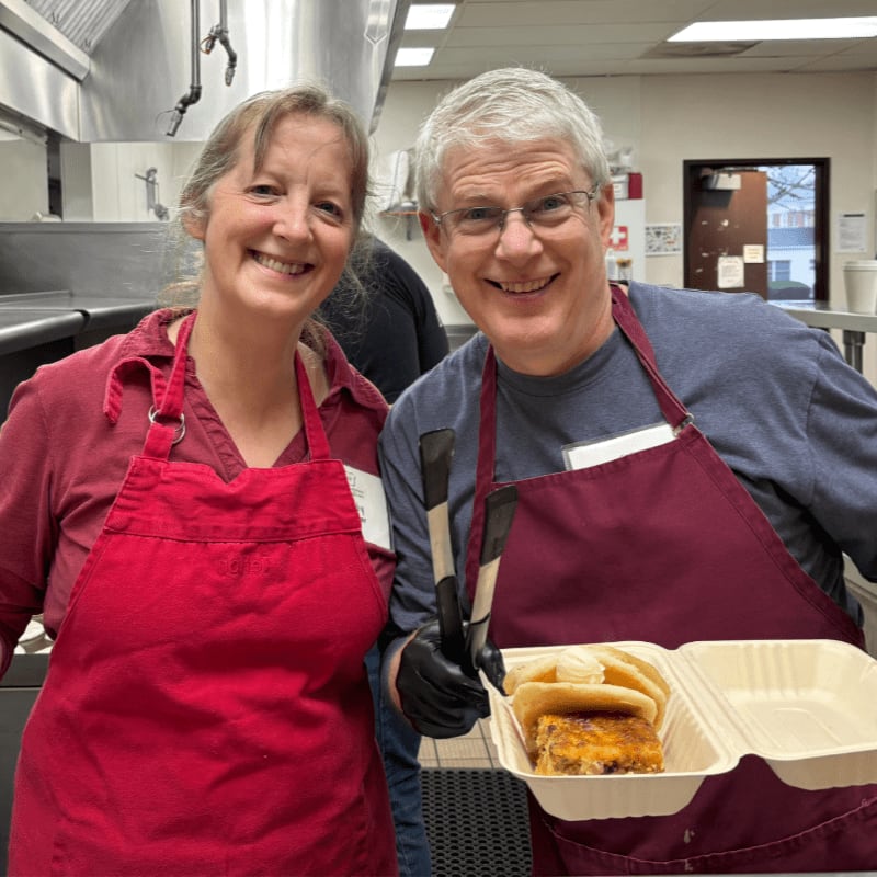 Two smiling volunteers in red aprons hold a takeout container of food in a commercial kitchen.