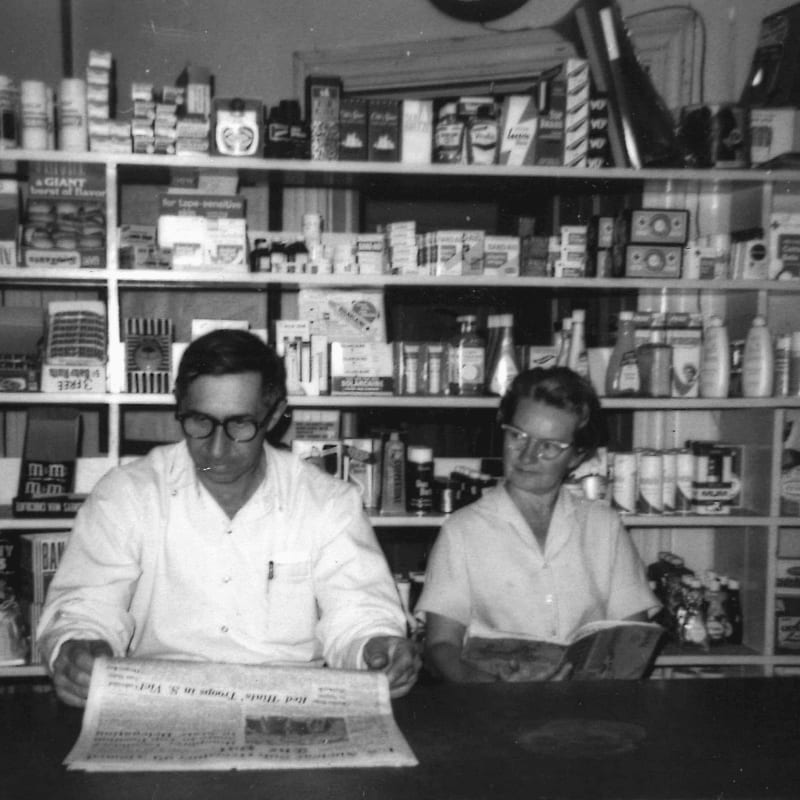 A man and a woman sit behind a counter in a store, reading, with shelves full of products in the background.