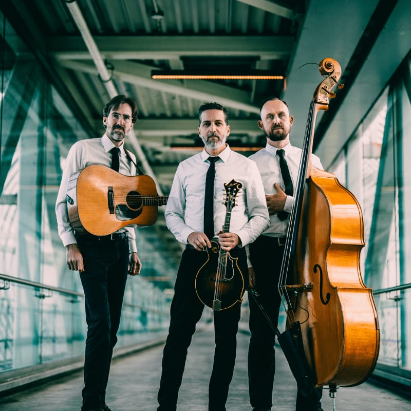 Three musicians in formal attire stand with their instruments in a modern, glass-enclosed walkway.