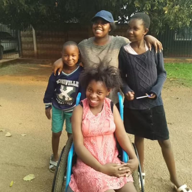 Four smiling children, one in a wheelchair, pose outdoors with a fence and trees in the background.