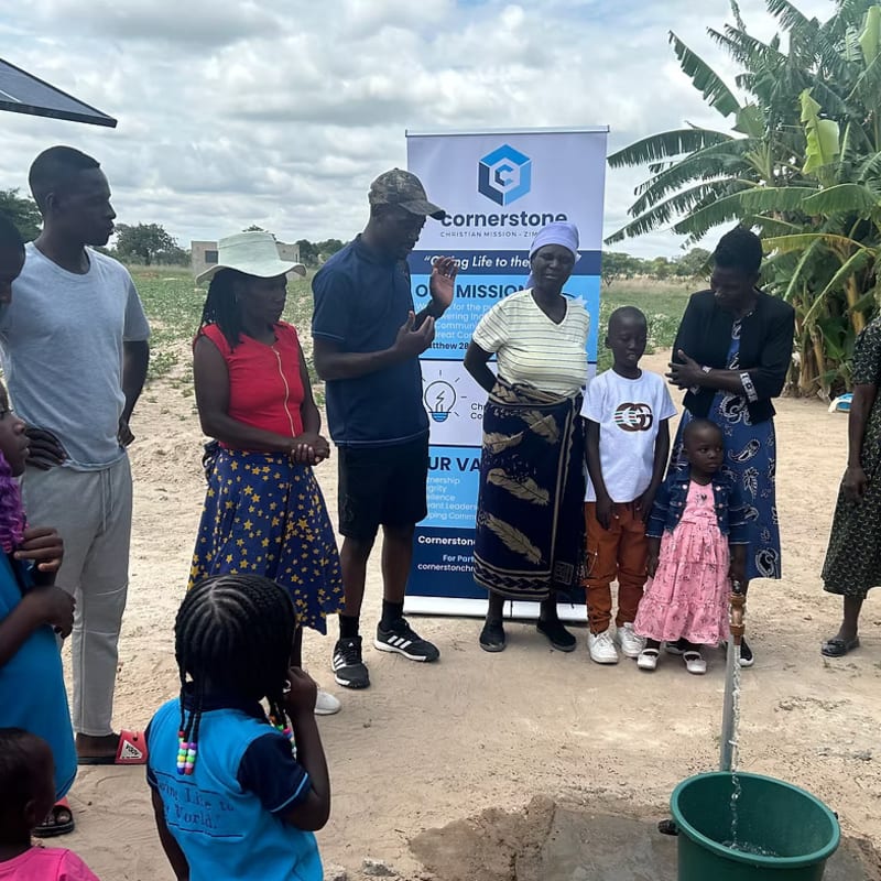 A group of people gather around a well in the foreground, with a Cornerstone banner and trees in the background.
