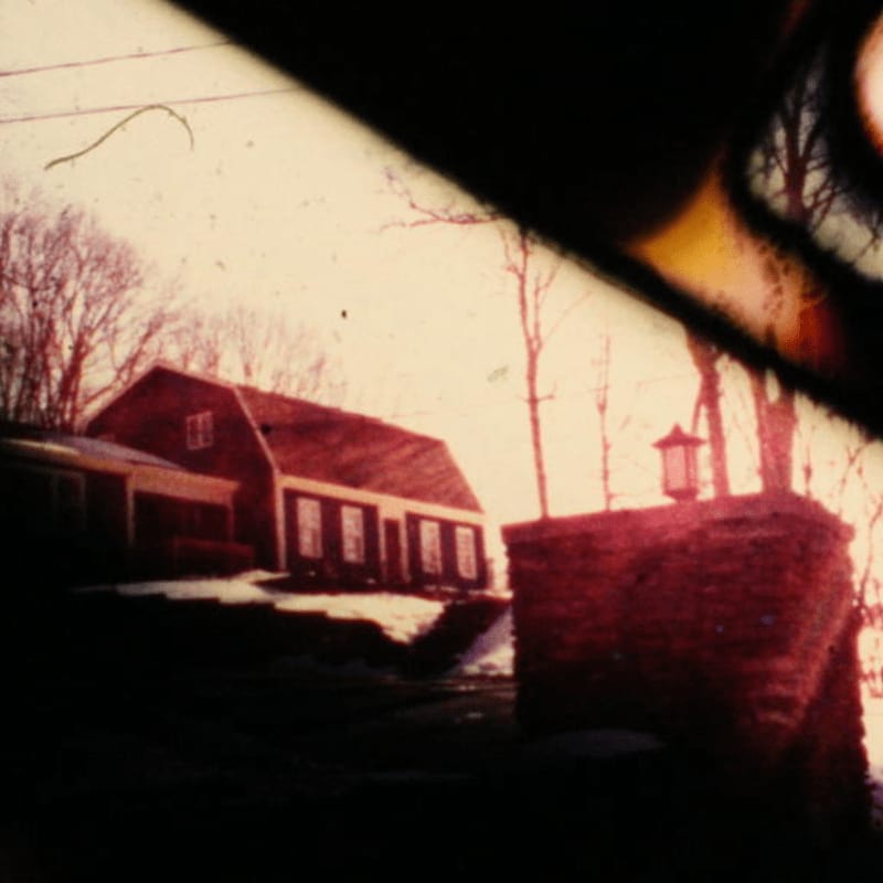 A red brick pillar with a lantern stands in the foreground, while a red house with snow-covered ground and bare trees fills the background.