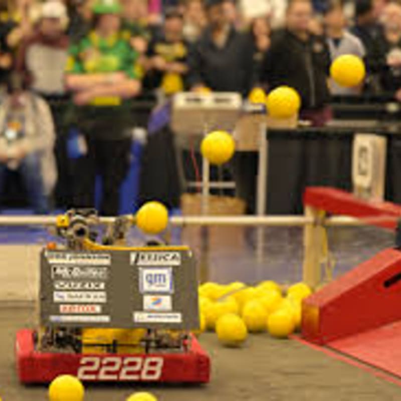 A red robot with the number 2228 on it sits on a dark floor in the foreground, while a crowd of people watches in the blurred background.