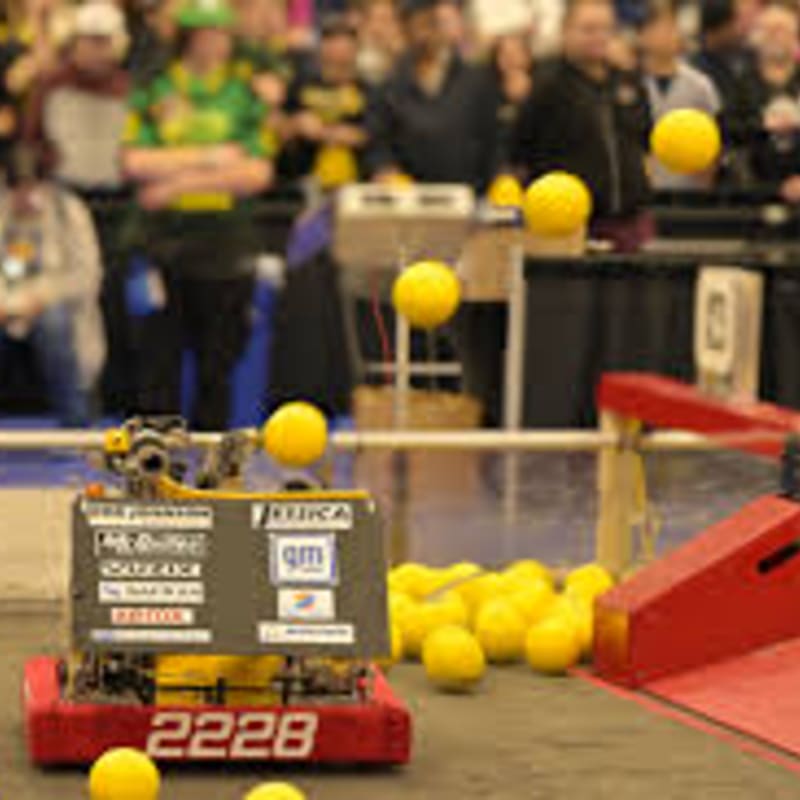 A red robot with the number 2228 on it is in the foreground, surrounded by yellow balls, while a blurred crowd watches in the background.