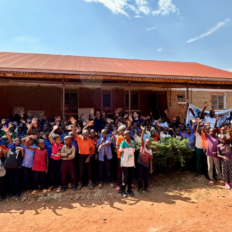 A large group of children wave enthusiastically in front of a red-roofed building under a clear blue sky.