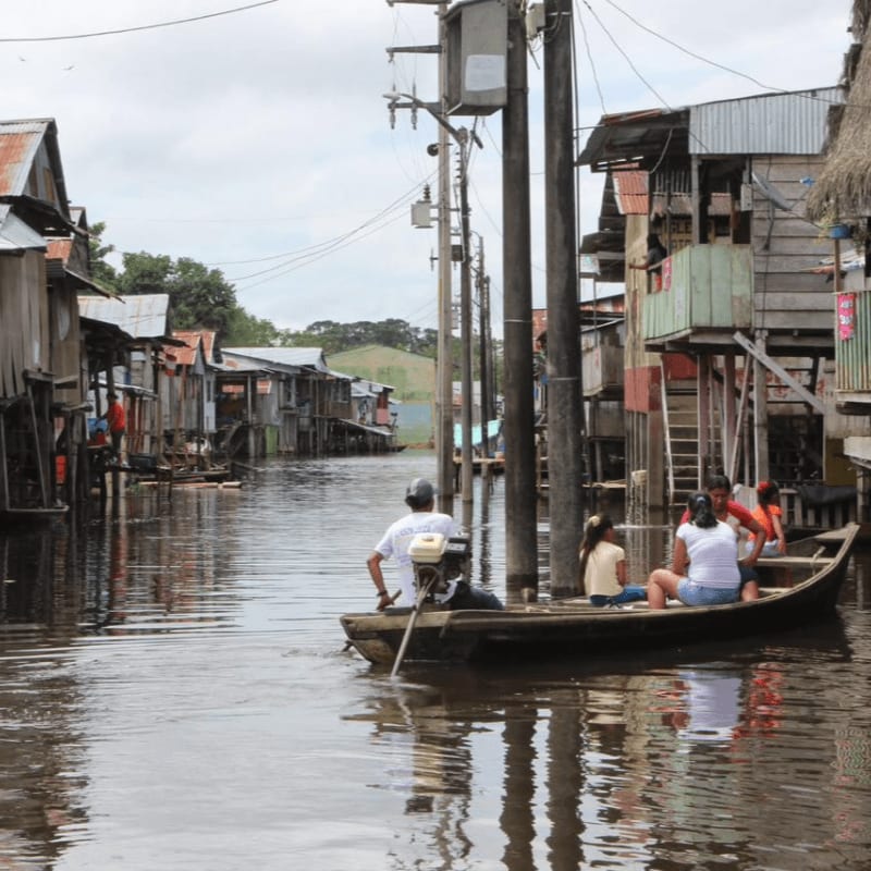 A small boat with people navigates through a flooded village with wooden houses and utility poles.