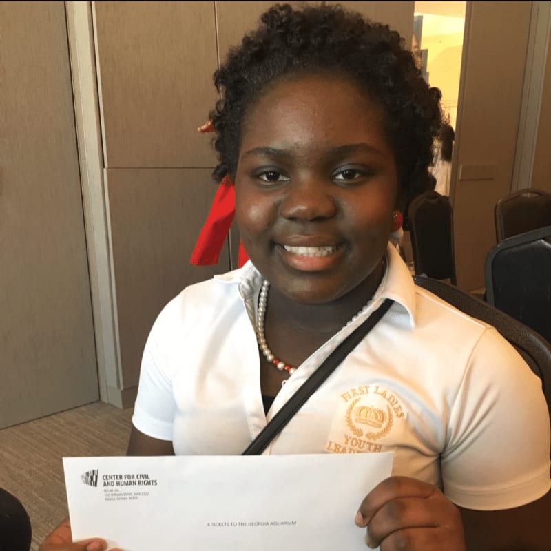 A young Black girl in a white polo shirt with a "First Ladies Youth Leadership" emblem smiles while holding a document in front of a blurred background of chairs and walls.