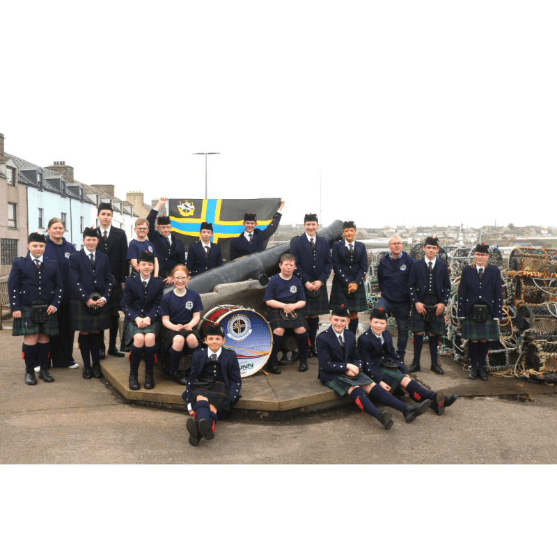 A group of people in kilts and uniforms pose around a cannon with a flag, while buildings and lobster pots form the background.