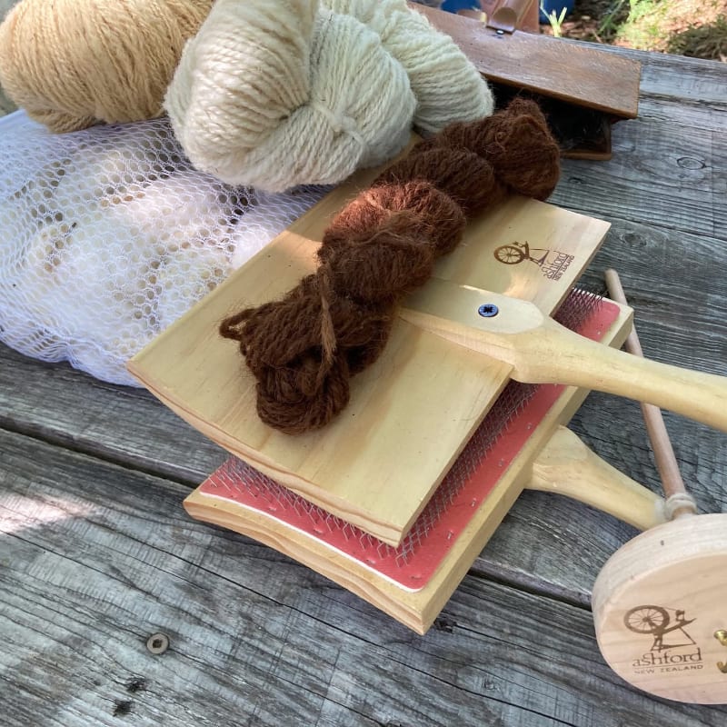 Brown yarn rests on wooden wool carders in the foreground, with balls of cream and tan yarn and a mesh bag of white fiber in the background.