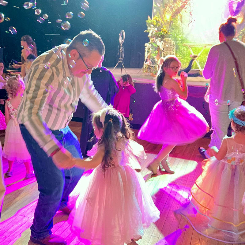 A man dances with a young girl in a pink dress in the foreground, while other children in similar attire and a decorated stage are visible in the background.