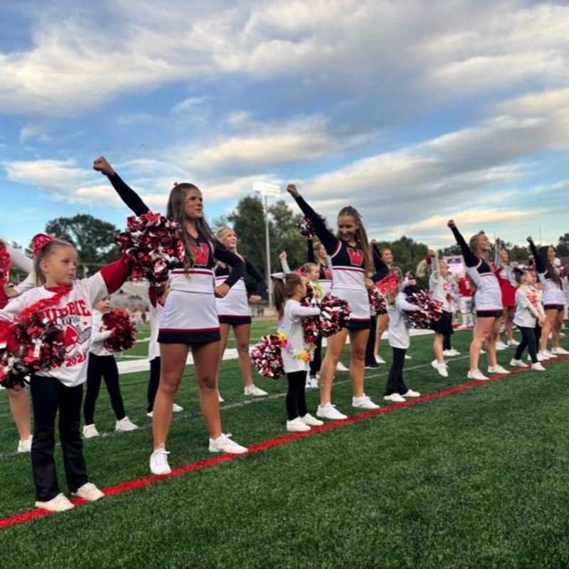 A group of young cheerleaders in uniform with red and white pom-poms stand on a green field with a cloudy sky in the background.