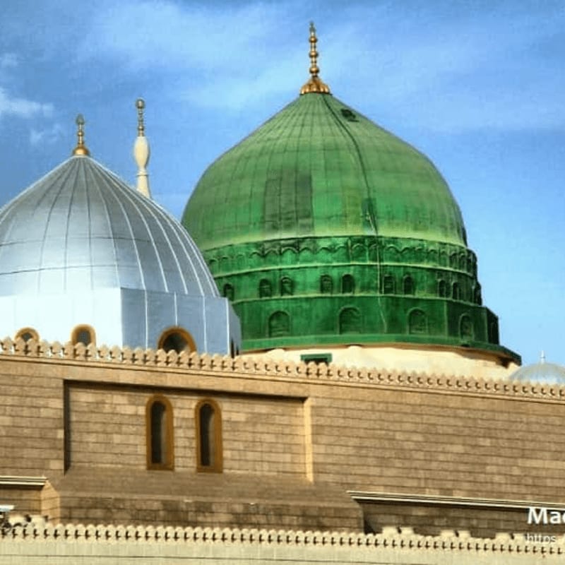 Two prominent domes, one green and one silver, rise above a stone building with arched windows, set against a bright blue sky with scattered clouds.