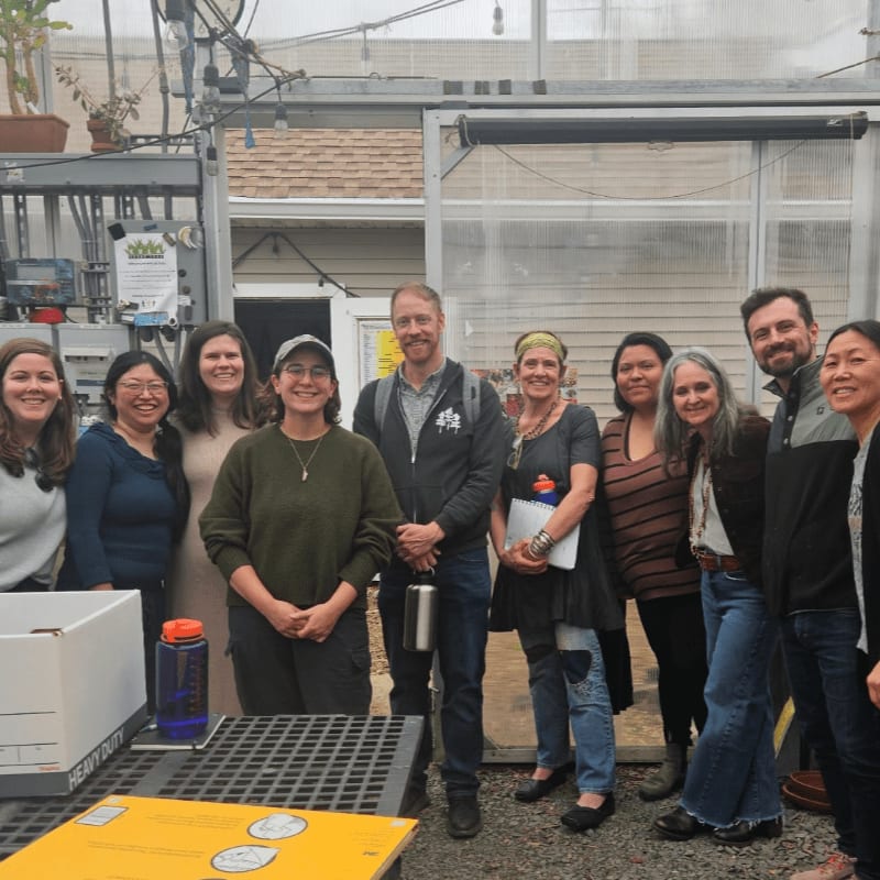 A group of people stand in front of a greenhouse, with a table in the foreground holding a box and water bottles.