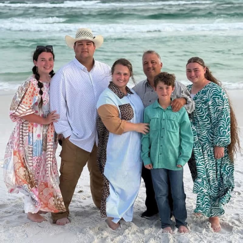A family of six poses on a sandy beach with the ocean waves in the background.