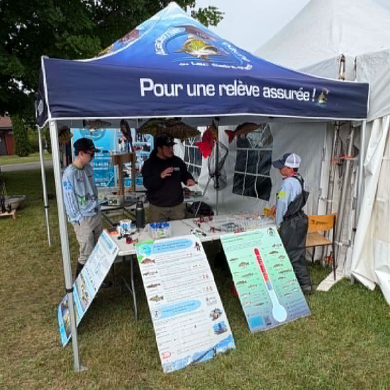 Three people stand behind a table displaying fishing gear and educational posters under a blue tent, with a white tent and trees visible in the background.